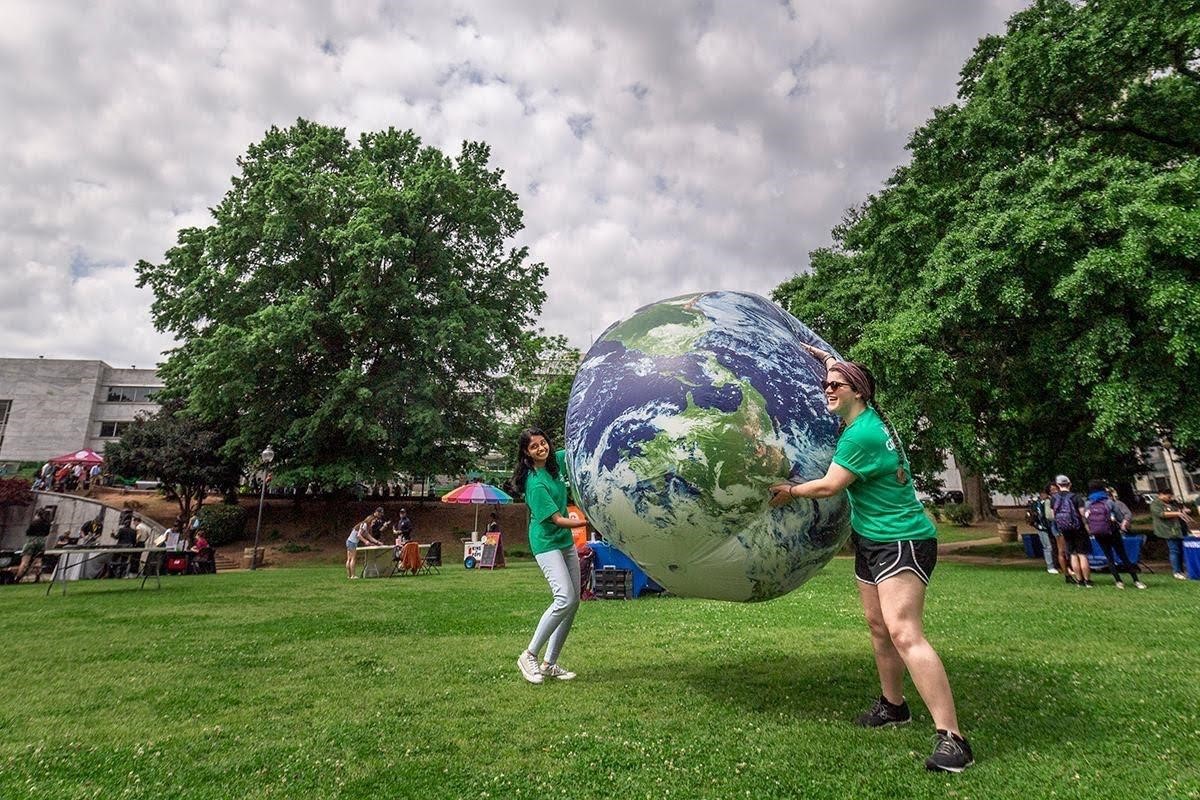 This is a picture of two women in green t-shirts holding an inflatable ball of the earth. They're outtside on grass. Trees with green leaves and a cloudy sky are in the background.