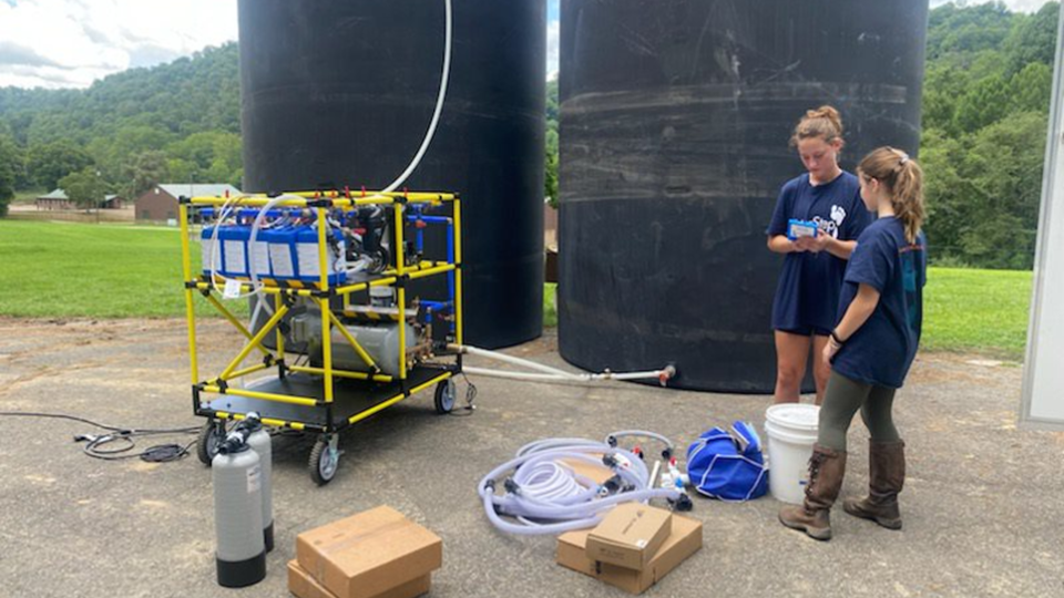 Two people look at a handheld water monitor. A WOW Cart is to the left and two black tanks stand behind them.